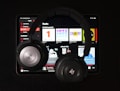 A pair of black over-ear headphones rests on top of a tablet displaying a music streaming app. Various music categories and album art are visible on the screen.