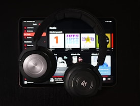 A pair of black over-ear headphones rests on top of a tablet displaying a music streaming app. Various music categories and album art are visible on the screen.
