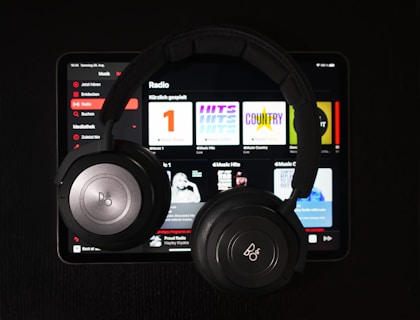 A pair of black over-ear headphones rests on top of a tablet displaying a music streaming app. Various music categories and album art are visible on the screen.