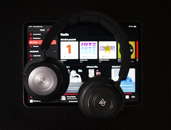 A pair of black over-ear headphones rests on top of a tablet displaying a music streaming app. Various music categories and album art are visible on the screen.