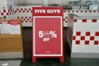 A promotional sign stands inside a restaurant, prominently displaying a 50% off discount. The sign is red with white text and is set in an interior with white and red checkered tiles. To the left, a poster detailing COVID-19 safety measures is visible.