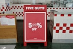 A promotional sign stands inside a restaurant, prominently displaying a 50% off discount. The sign is red with white text and is set in an interior with white and red checkered tiles. To the left, a poster detailing COVID-19 safety measures is visible.
