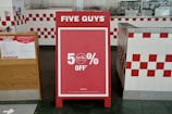 A promotional sign stands inside a restaurant, prominently displaying a 50% off discount. The sign is red with white text and is set in an interior with white and red checkered tiles. To the left, a poster detailing COVID-19 safety measures is visible.