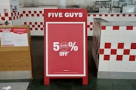 A promotional sign stands inside a restaurant, prominently displaying a 50% off discount. The sign is red with white text and is set in an interior with white and red checkered tiles. To the left, a poster detailing COVID-19 safety measures is visible.