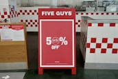 A promotional sign stands inside a restaurant, prominently displaying a 50% off discount. The sign is red with white text and is set in an interior with white and red checkered tiles. To the left, a poster detailing COVID-19 safety measures is visible.