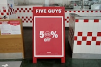 A promotional sign stands inside a restaurant, prominently displaying a 50% off discount. The sign is red with white text and is set in an interior with white and red checkered tiles. To the left, a poster detailing COVID-19 safety measures is visible.
