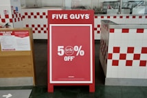 A promotional sign stands inside a restaurant, prominently displaying a 50% off discount. The sign is red with white text and is set in an interior with white and red checkered tiles. To the left, a poster detailing COVID-19 safety measures is visible.