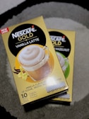 Two boxes of Nescafé Gold coffee are displayed on a gray surface. The boxes are yellow and feature images of coffee in clear mugs with frothy tops. One box advertises 'Vanilla Latte' flavored coffee, while the other partially visible box is for 'Hazelnut' flavored coffee. The boxes also indicate that each contains 10 servings.