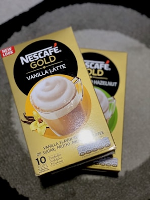 Two boxes of Nescafé Gold coffee are displayed on a gray surface. The boxes are yellow and feature images of coffee in clear mugs with frothy tops. One box advertises 'Vanilla Latte' flavored coffee, while the other partially visible box is for 'Hazelnut' flavored coffee. The boxes also indicate that each contains 10 servings.
