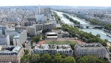 An aerial view of a large city with a prominent river running through it. The urban landscape is densely packed with various architectural styles, including modern high-rises and older residential buildings. Bridges span the river, connecting different parts of the city, and a sports field is visible surrounded by greenery.