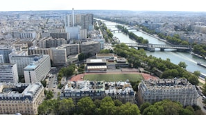 An aerial view of a large city with a prominent river running through it. The urban landscape is densely packed with various architectural styles, including modern high-rises and older residential buildings. Bridges span the river, connecting different parts of the city, and a sports field is visible surrounded by greenery.