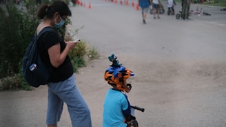A woman wearing a face mask and holding a phone stands next to a child with a bicycle helmet, who is on a balance bike. Orange traffic cones are lined up in the background, along with other people and children participating in outdoor activities.