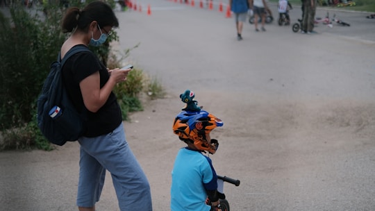 A woman wearing a face mask and holding a phone stands next to a child with a bicycle helmet, who is on a balance bike. Orange traffic cones are lined up in the background, along with other people and children participating in outdoor activities.