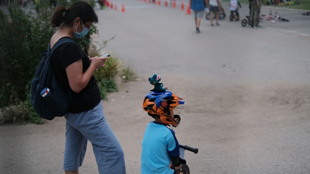 A woman wearing a face mask and holding a phone stands next to a child with a bicycle helmet, who is on a balance bike. Orange traffic cones are lined up in the background, along with other people and children participating in outdoor activities.
