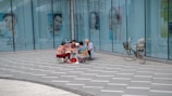 A group of people are seated on small folding chairs, engaged in an activity together on a paved sidewalk outside a large glass building displaying advertisements with various faces. A bicycle is parked nearby, leaning against the building's wall.
