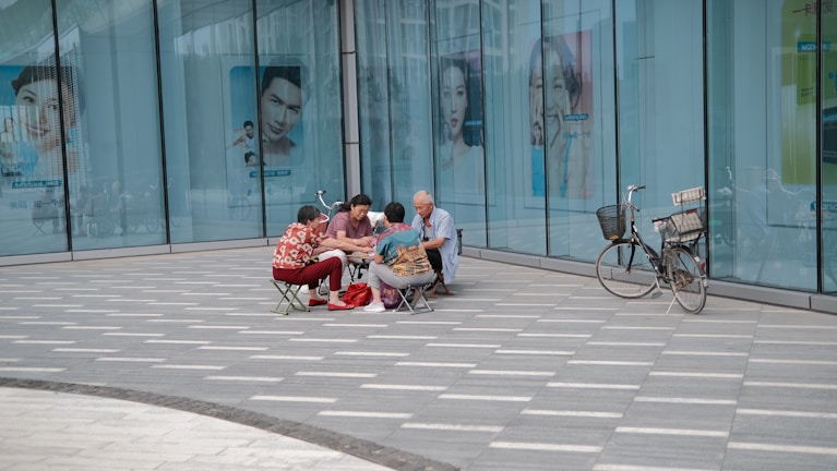 A group of people are seated on small folding chairs, engaged in an activity together on a paved sidewalk outside a large glass building displaying advertisements with various faces. A bicycle is parked nearby, leaning against the building's wall.