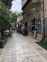 A narrow paved street lined with stone buildings and trees, where people are casually sitting at outdoor tables. Signs for a travel agency are prominently displayed, along with various travel posters. The atmosphere is relaxed and inviting, with greenery providing contrast to the stone structures.