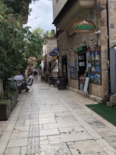 A narrow paved street lined with stone buildings and trees, where people are casually sitting at outdoor tables. Signs for a travel agency are prominently displayed, along with various travel posters. The atmosphere is relaxed and inviting, with greenery providing contrast to the stone structures.