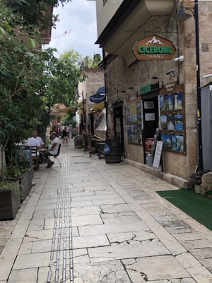A narrow paved street lined with stone buildings and trees, where people are casually sitting at outdoor tables. Signs for a travel agency are prominently displayed, along with various travel posters. The atmosphere is relaxed and inviting, with greenery providing contrast to the stone structures.