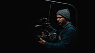 A person wearing a beanie is holding a professional video camera in a dark setting. The camera appears to be mounted on a stabilization rig, suggesting a focus on film-making or videography.