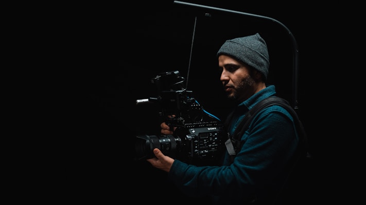 A person wearing a beanie is holding a professional video camera in a dark setting. The camera appears to be mounted on a stabilization rig, suggesting a focus on film-making or videography.
