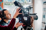 A person wearing headphones is adjusting a professional video camera setup, which includes a large lens and various attachments. The background features blurred, colorful patterns and soft natural light from a window.