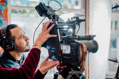 A person wearing headphones is adjusting a professional video camera setup, which includes a large lens and various attachments. The background features blurred, colorful patterns and soft natural light from a window.