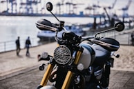 A sleek motorcycle with gold front forks and a round headlamp is parked near a waterfront. The background includes blurred images of people walking and an industrial harbor with cranes and shipping containers in the distance.