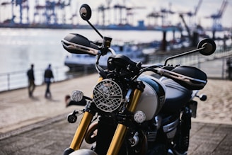 A sleek motorcycle with gold front forks and a round headlamp is parked near a waterfront. The background includes blurred images of people walking and an industrial harbor with cranes and shipping containers in the distance.