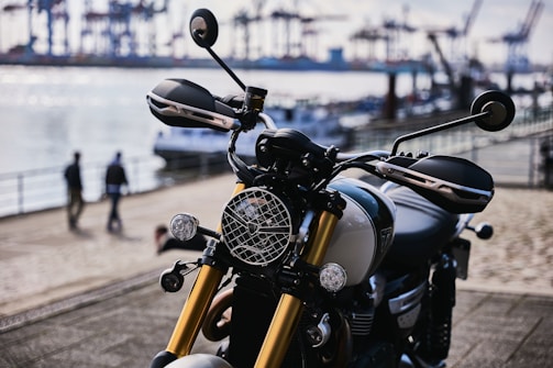 A sleek motorcycle with gold front forks and a round headlamp is parked near a waterfront. The background includes blurred images of people walking and an industrial harbor with cranes and shipping containers in the distance.