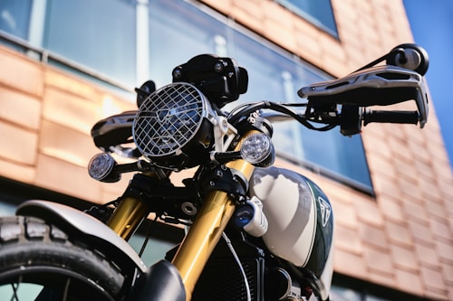 A close-up view of a motorcycle with an angular building in the background. The motorcycle features a large circular headlight with a protective grill, two smaller round lights, and sturdy handlebars. The design on the fuel tank is sleek, and the front suspension is highlighted with gold-colored components.