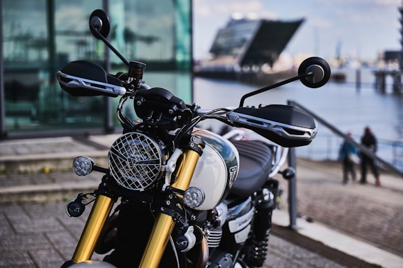 A close-up view of a modern motorcycle with a focus on the handlebars and front suspension. Behind the motorcycle, there is an urban waterfront scene with a geometric-shaped building and bodies of water. The background also shows people walking near the edge of the water.
