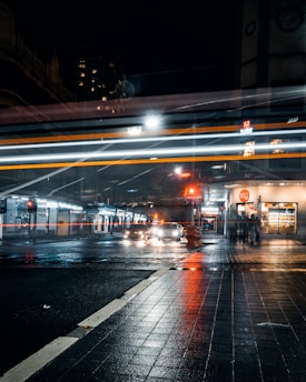 A nighttime urban street scene with blurred motion effects of cars and city lights. The wet pavement reflects the glow of headlights and street lamps, creating an atmospheric cityscape. A storefront is visible with a few pedestrians moving along the sidewalk.