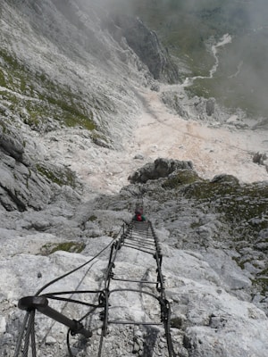 A steep rocky mountain terrain with a metal ladder extending downwards. A person in climbing gear is ascending or descending the ladder. Surrounding the ladder, rough and jagged rocks dominate the scene with a sparse covering of greenery in patches.