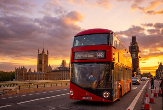 A red double-decker bus is driving on a bridge in London with the iconic Big Ben and the Houses of Parliament in the background. The sky is filled with vibrant colors from the setting sun, casting a warm glow over the scene. Traffic cones and a few people are visible on the road.