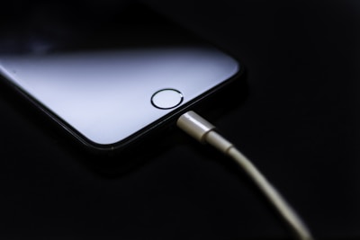 A smartphone with a visible home button lies on a dark surface, connected to a charging cable. The lighting highlights the sleek design of the phone while the cable is plugged into the phone's charging port.