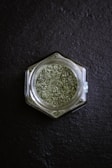 A hexagonal glass jar filled with green dried herbs is placed on a dark slate surface. The finely chopped herbs create a textured appearance, and the clear glass of the jar reflects some light.