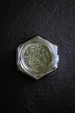 A hexagonal glass jar filled with green dried herbs is placed on a dark slate surface. The finely chopped herbs create a textured appearance, and the clear glass of the jar reflects some light.