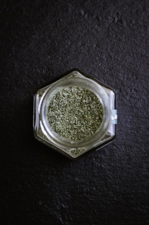 A hexagonal glass jar filled with green dried herbs is placed on a dark slate surface. The finely chopped herbs create a textured appearance, and the clear glass of the jar reflects some light.