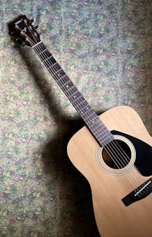 An acoustic guitar is positioned against a floral-patterned wall. The guitar features a natural wood body with a glossy finish and a dark fretboard with metal frets. The headstock displays tuning pegs and a brand label.