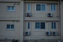 A building facade featuring several windows and multiple air conditioning units mounted on the walls. The walls are beige and slightly weathered, with visible pipes running vertically and horizontally. There are small external lamps and some foliage visible at the bottom.