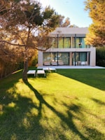 A modern two-story house with large glass windows and a spacious backyard. The house is surrounded by trees, and the large lawn is well-manicured with the shadow of a tree covering part of the grass. There are outdoor furniture pieces including lounge chairs and a table with chairs on the patio area near the house.