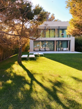 A modern two-story house with large glass windows and a spacious backyard. The house is surrounded by trees, and the large lawn is well-manicured with the shadow of a tree covering part of the grass. There are outdoor furniture pieces including lounge chairs and a table with chairs on the patio area near the house.