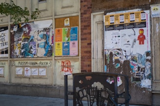 A weathered urban wall covered with a variety of overlapping posters and graffiti. The posters include calls to shop locally, political slogans, and references to Marxism-Leninism-Maoism. There's a mix of vibrant and faded colors. Some posters are partially torn, and there's a black metal fence in the foreground.