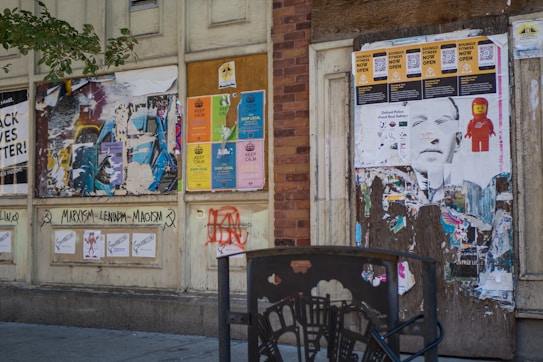 A weathered urban wall covered with a variety of overlapping posters and graffiti. The posters include calls to shop locally, political slogans, and references to Marxism-Leninism-Maoism. There's a mix of vibrant and faded colors. Some posters are partially torn, and there's a black metal fence in the foreground.