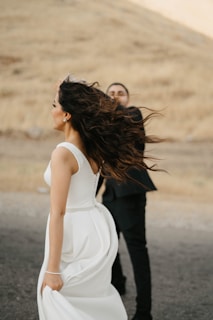 A bride in a white gown appears to be spinning in an outdoor setting, with long hair flowing. A groom in a dark suit stands partially obscured behind her. The background features a dry, grassy hill under soft, diffused lighting.