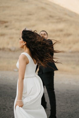 A bride in a white gown appears to be spinning in an outdoor setting, with long hair flowing. A groom in a dark suit stands partially obscured behind her. The background features a dry, grassy hill under soft, diffused lighting.