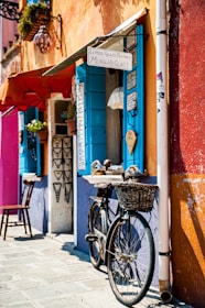 A quaint street scene featuring a bicycle with a wicker basket leaning against a colorful building. The building is adorned with vibrant shades of orange, blue, and purple, and displays a sign for a Murano glass shop. Potted plants and decorative necklaces hang near the window, which has bright blue shutters. A wooden chair is placed on the stone pavement.