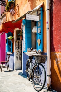 A quaint street scene featuring a bicycle with a wicker basket leaning against a colorful building. The building is adorned with vibrant shades of orange, blue, and purple, and displays a sign for a Murano glass shop. Potted plants and decorative necklaces hang near the window, which has bright blue shutters. A wooden chair is placed on the stone pavement.