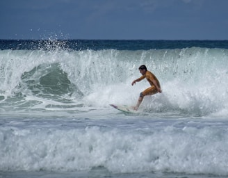 A surfer skillfully rides a wave, maintaining balance and focus. The ocean wave is large and crashing, with white foam spraying around. The surfer is wearing a bright yellow wetsuit, standing out against the blue-green sea.
